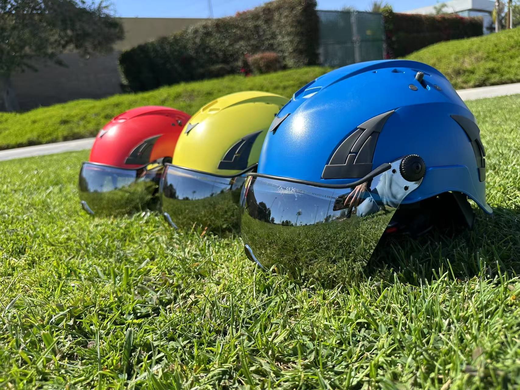 Low angle of three AeroPro Safety Helmets with Smoke Visor Kit, colors yellow, red and blue on Grass. Three ANSI/ISEA Z89.1-2014 Type 1 Class C Safety Helmets with Smoke Visor used for Industrial, Rescue & fall protection.