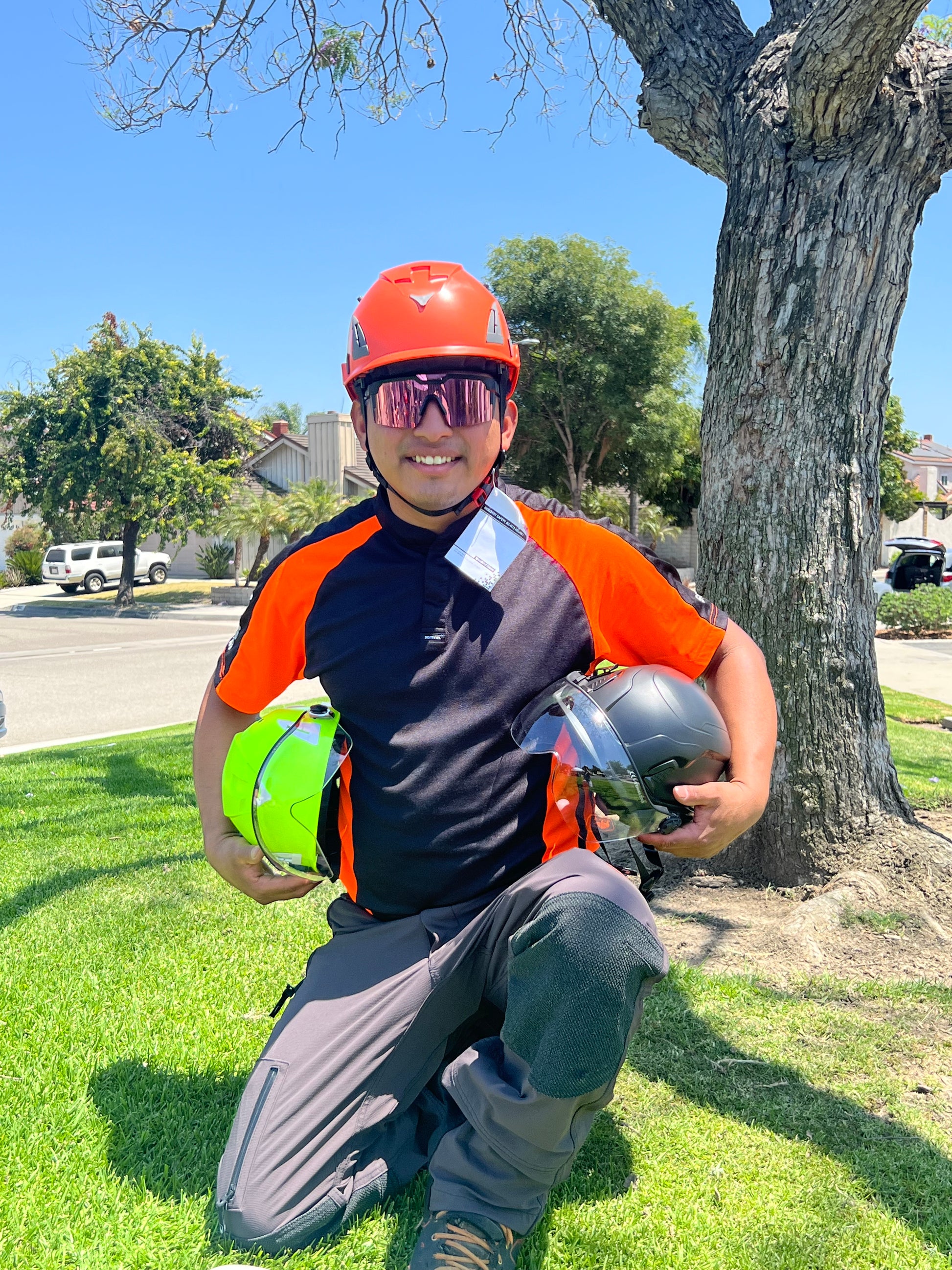 Front view of construction worker with orange AeroPro Safety Helmet ANSI/ISEA Z89.1-2014 OSHA Certified Type 1 Class C on head kneeling on grass, while holding orange and black color variations with clear visor.