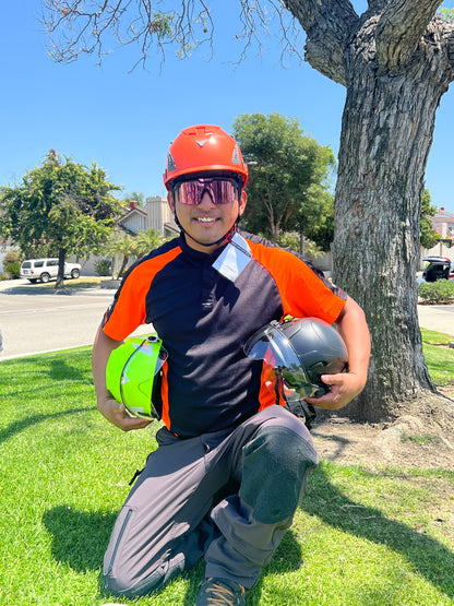 Front view of construction worker with orange AeroPro Safety Helmet ANSI/ISEA Z89.1-2014 OSHA Certified Type 1 Class C on head kneeling on grass, while holding orange and black color variations with clear visor.
